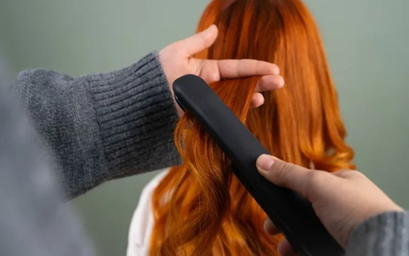 Hands styling long red hair using a flat iron indoors. Haircare and beauty concept.