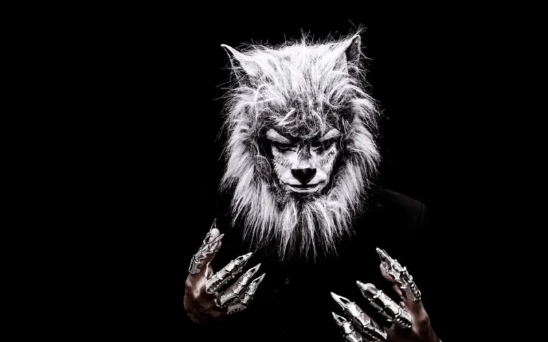 Striking portrait of an adult in a realistic werewolf costume with metallic claws against a black background.