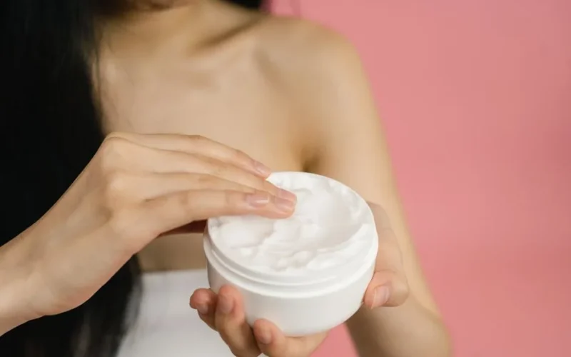 Close-up of a woman applying white cream from a jar against a pink background.
