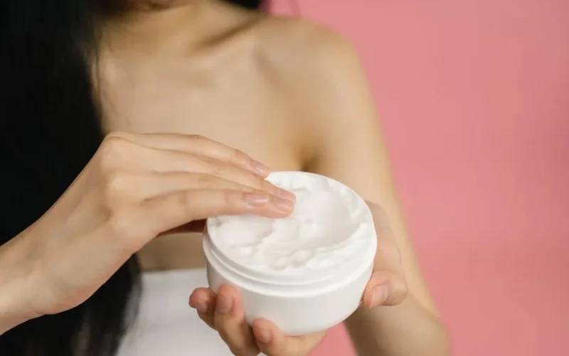 Close-up of a woman applying white cream from a jar against a pink background.