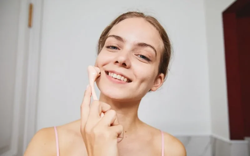 Happy woman enjoying a skincare routine using a facial massage roller indoors.