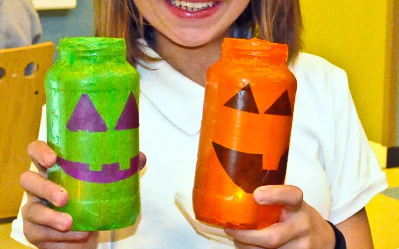 Thalia Schober, a seventh-grader from Ross Middle School holds up two Jar-O-Lanterns: decorative glass mason jars that resemble Halloween pumpkins and other Halloween decor. When you light the candle on the inside of the jar it gives off a colorful glow that brings forth a spooky festive ambience that she made during the Spooky Arts and Crafts activity.