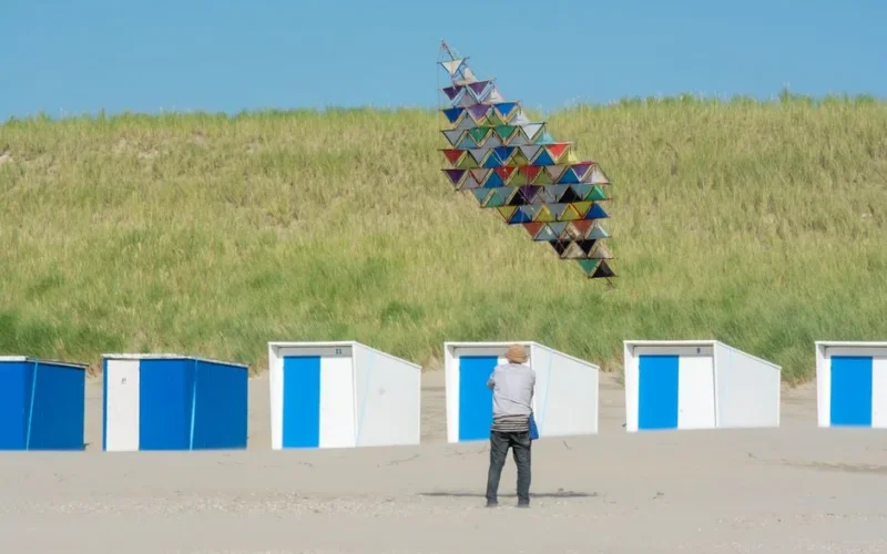 A man flies a vibrant kite near blue and white beach cabins on a sunny day.