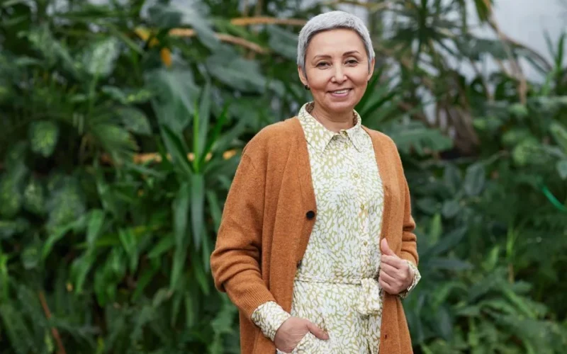 Portrait of a joyful senior woman in a stylish cardigan and dress, smiling in a vibrant greenhouse setting.