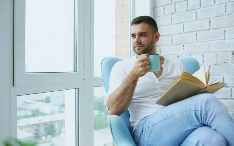 Man enjoys a quiet moment with coffee and a book in a cozy chair by the window.