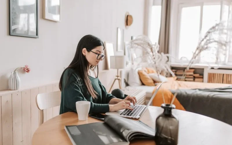 Side view of female remote worker in eyeglasses and casual wear typing on netbook while sitting at wooden table with cup and open magazine in bedroom at home