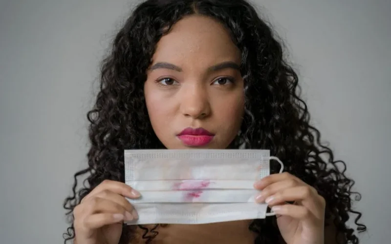 A woman with short blonde hair adjusts her black face mask indoors for safety.