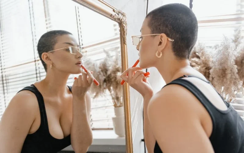 A woman in glasses applies lipstick while looking in the mirror. Bright, modern, and stylish.