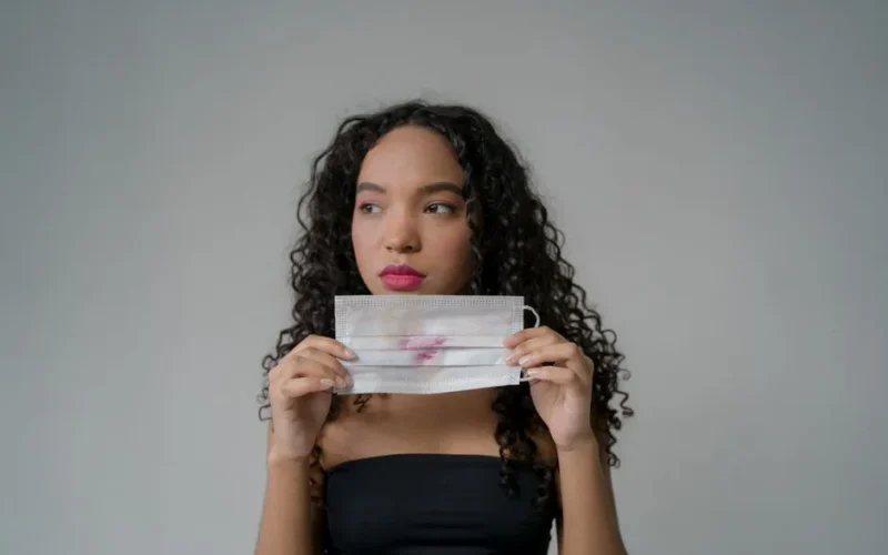 Young woman with curly hair holding a mask marked by lipstick, reflecting on post-pandemic life.