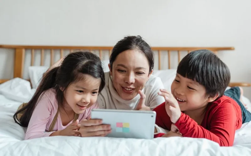 Full body smiling Asian mother and kids in casual clothes lying on stomachs on comfortable bed while spending time together playing game via tablet