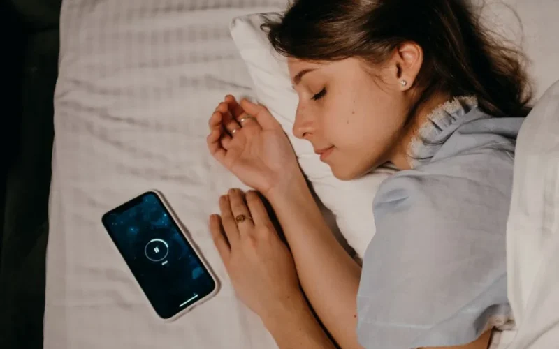 A woman peacefully sleeps in bed with a smartphone beside her, illustrating modern bedtime habits.