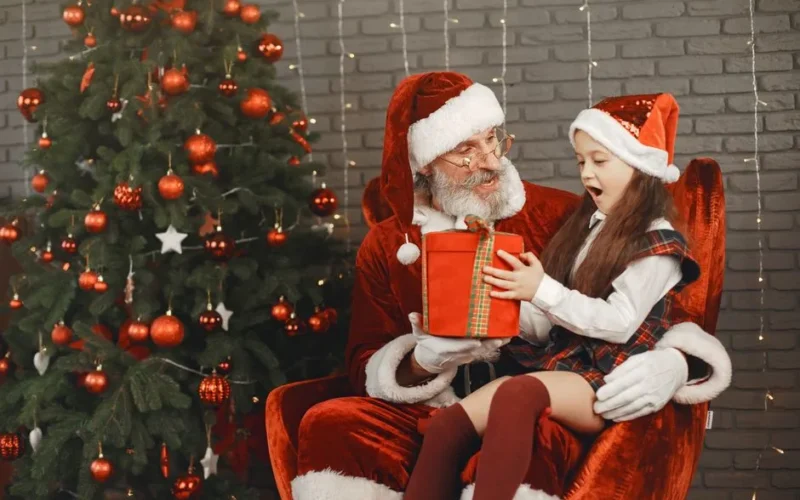 Santa Claus surprises a little girl with a Christmas gift by the tree indoors. Joyful holiday moment.