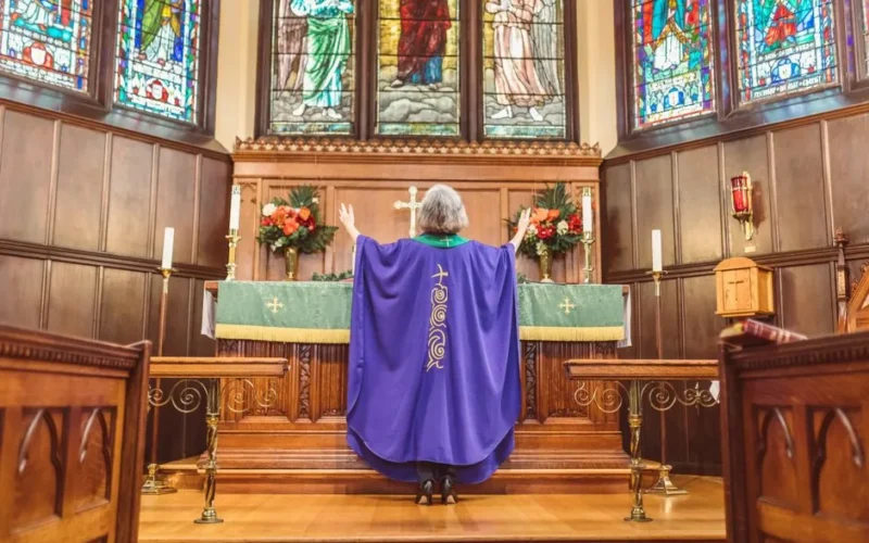 A priest stands at the altar in a church, leading prayer with arms raised, surrounded by stained glass windows.