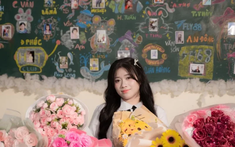 A young woman with bouquets of flowers stands in front of a decorated classroom chalkboard.