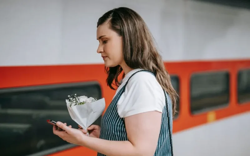A woman stands at a train station holding flowers and using her phone.