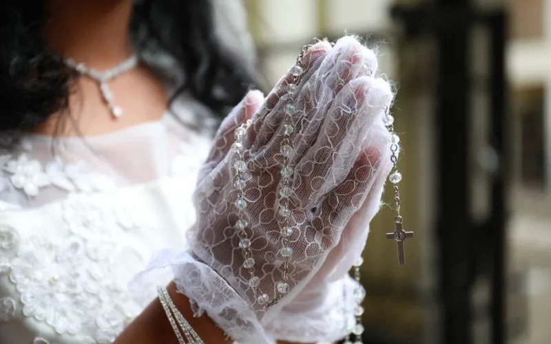 Hands in prayer with lace gloves holding rosary beads, symbolizing a First Communion ceremony.