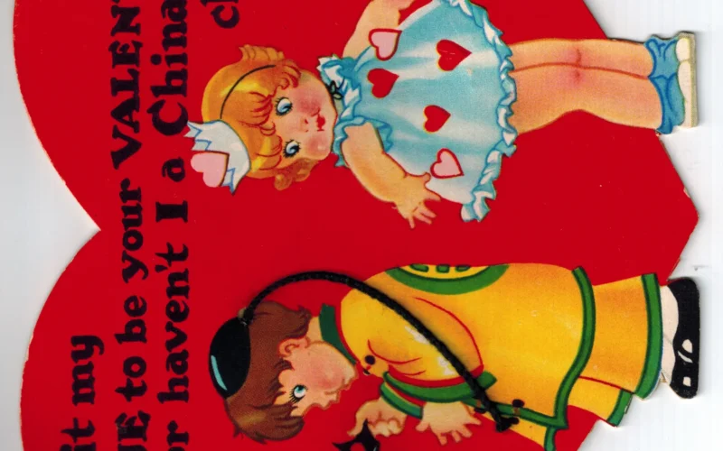 Valentine's Day card for children, which is captioned "Is it my CUE to be your VALENTINE or haven't I a Chinaman's chance?" Based on the art style, this appears to date back to the 1930s - 1950s. The boy, on the left, has a cap with a long string, referring to the queue worn by Chinese men during the Manchu dynasty (1644-1911). The girl, on the right, is wearing a blue babydoll dress with red and pink hearts, and is looking at the boy.