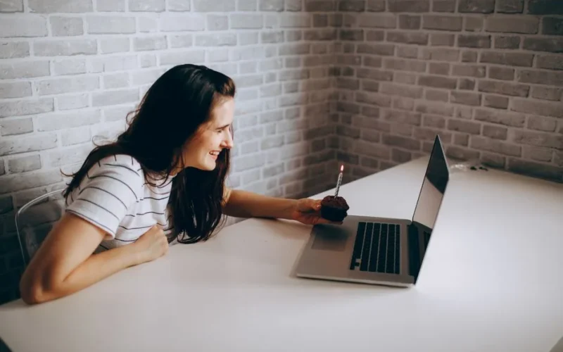 Woman smiling at laptop during a birthday video call, holding a cupcake with a lit candle.