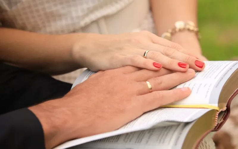 Close-up of a couple with wedding rings, hands resting on an open Bible, symbolizing love and faith.