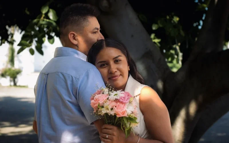 A couple lovingly embraces under a tree, holding a bouquet of pink and white roses.