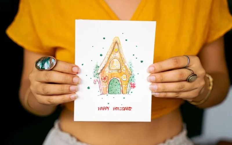 Woman holding a handmade holiday card with a gingerbread house illustration.