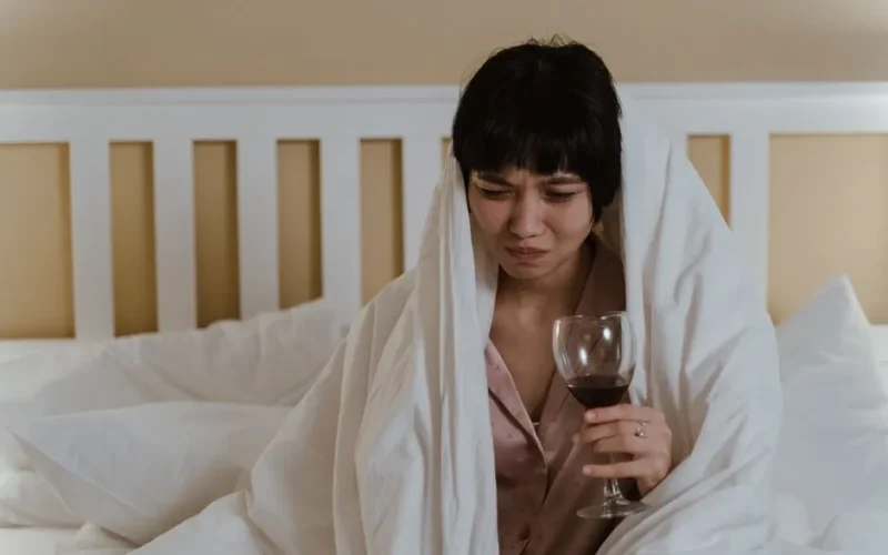 A woman wrapped in a blanket holding a wine glass, showing a look of discomfort.