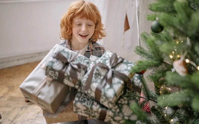 A smiling boy holds wrapped Christmas gifts indoors by a tree, celebrating holiday joy.