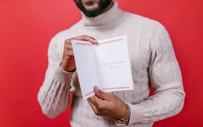A man in a sweater holds a Valentine's card on a red background.