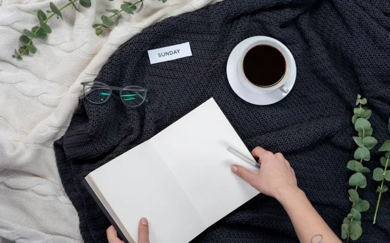 A cozy setup with black coffee, notebook, glasses, and a Sunday note on a knitted blanket.