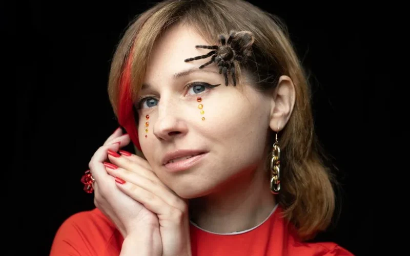Bizarre young female with big hairy spider on face in Halloween costume against black background