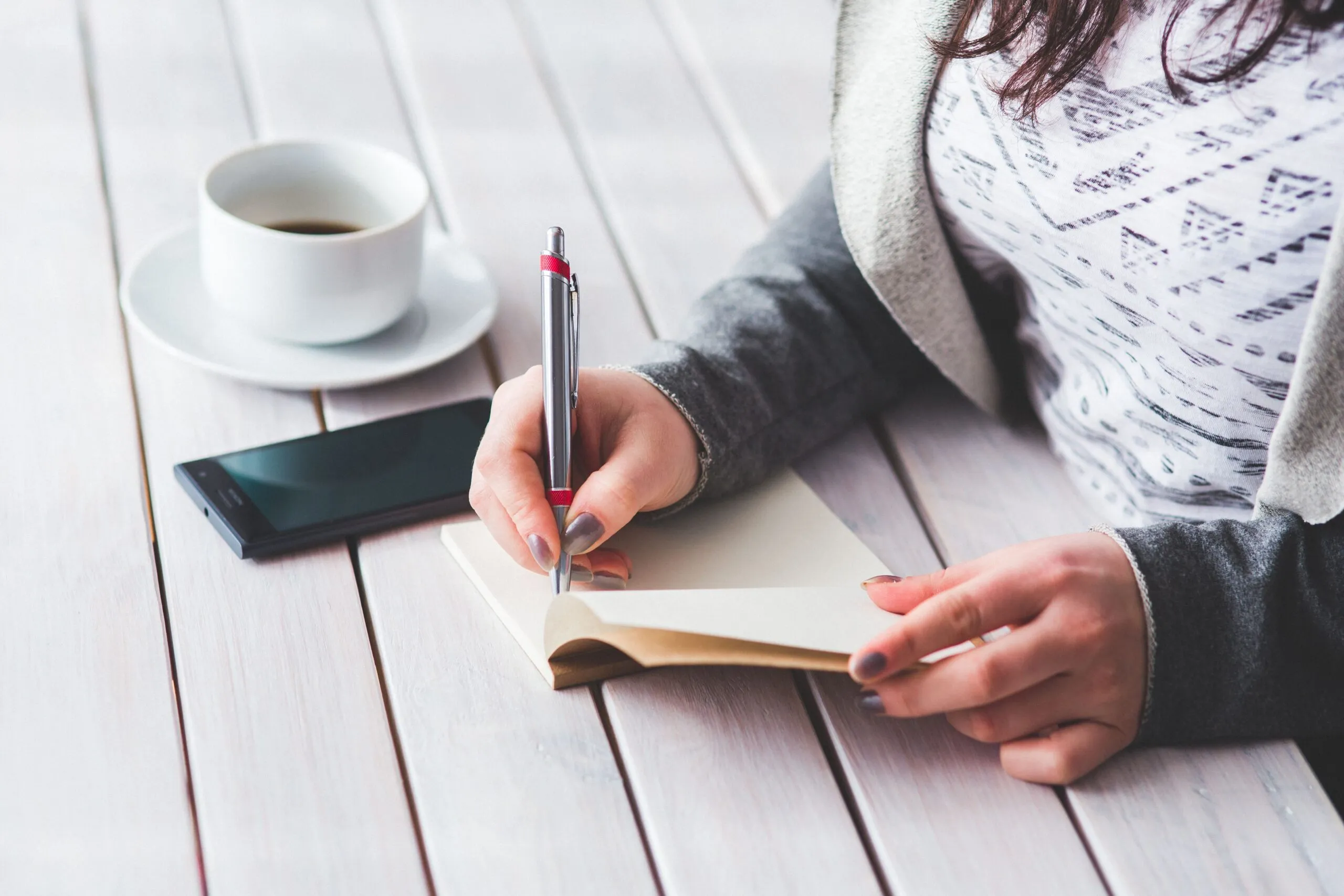 A woman writes in a journal next to a cup of coffee and a smartphone on a wooden table.