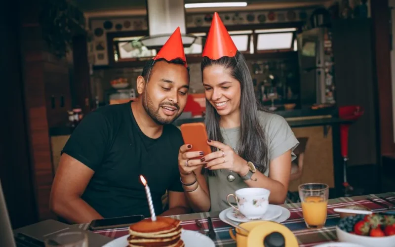 Two friends enjoying a birthday brunch indoors, wearing party hats and using a smartphone.