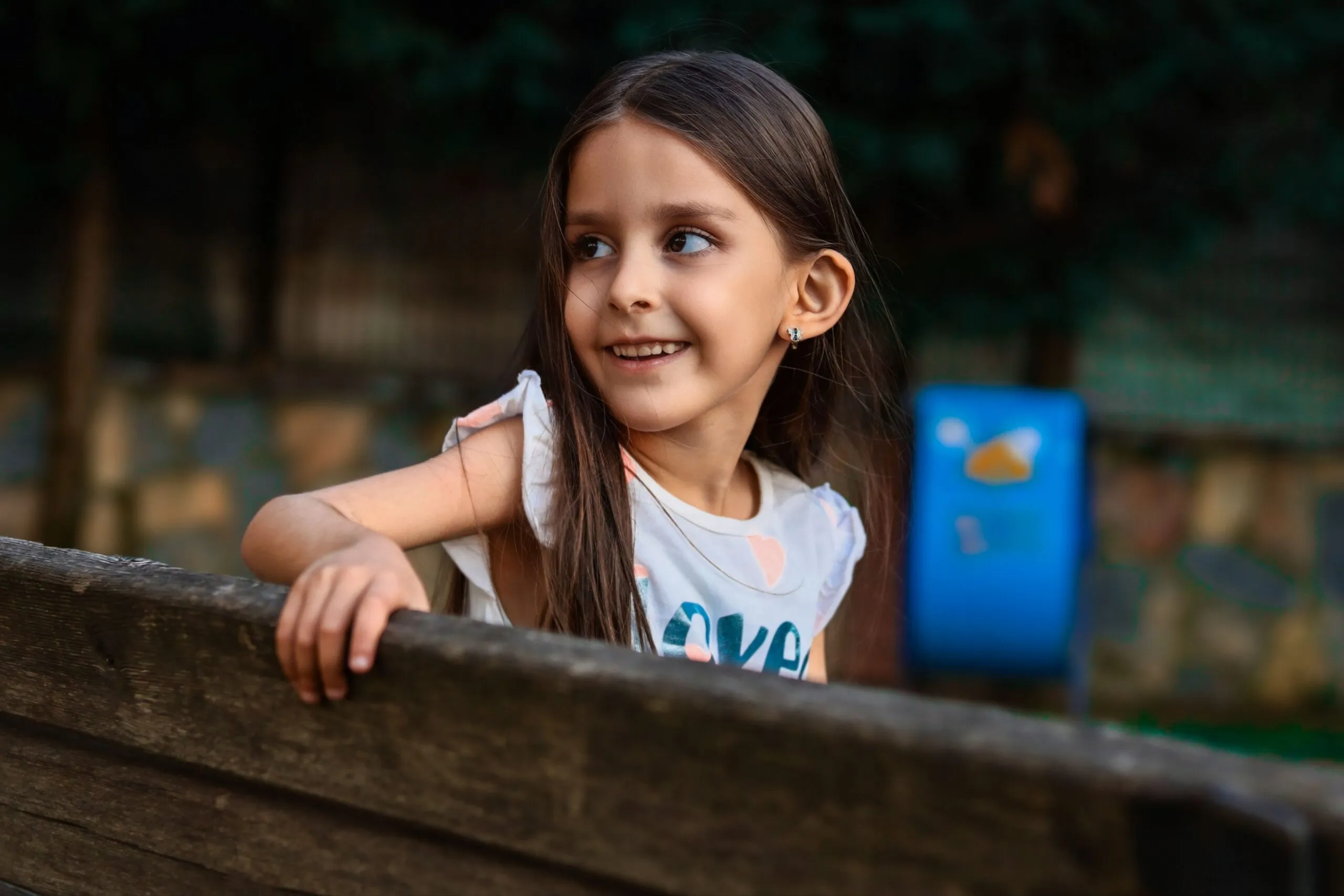 Young girl smiling while leaning on a wooden bench outdoors in a park setting.