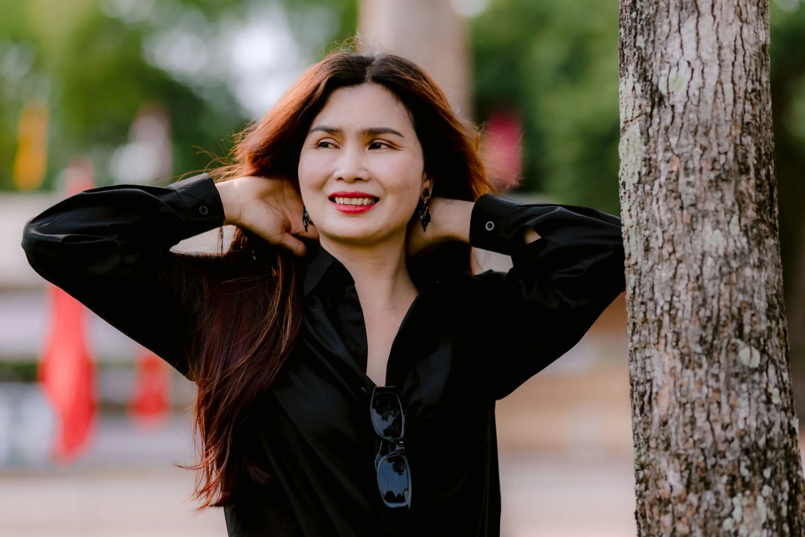 A woman smiling and relaxing outdoors, leaning against a tree in a sunny park setting.