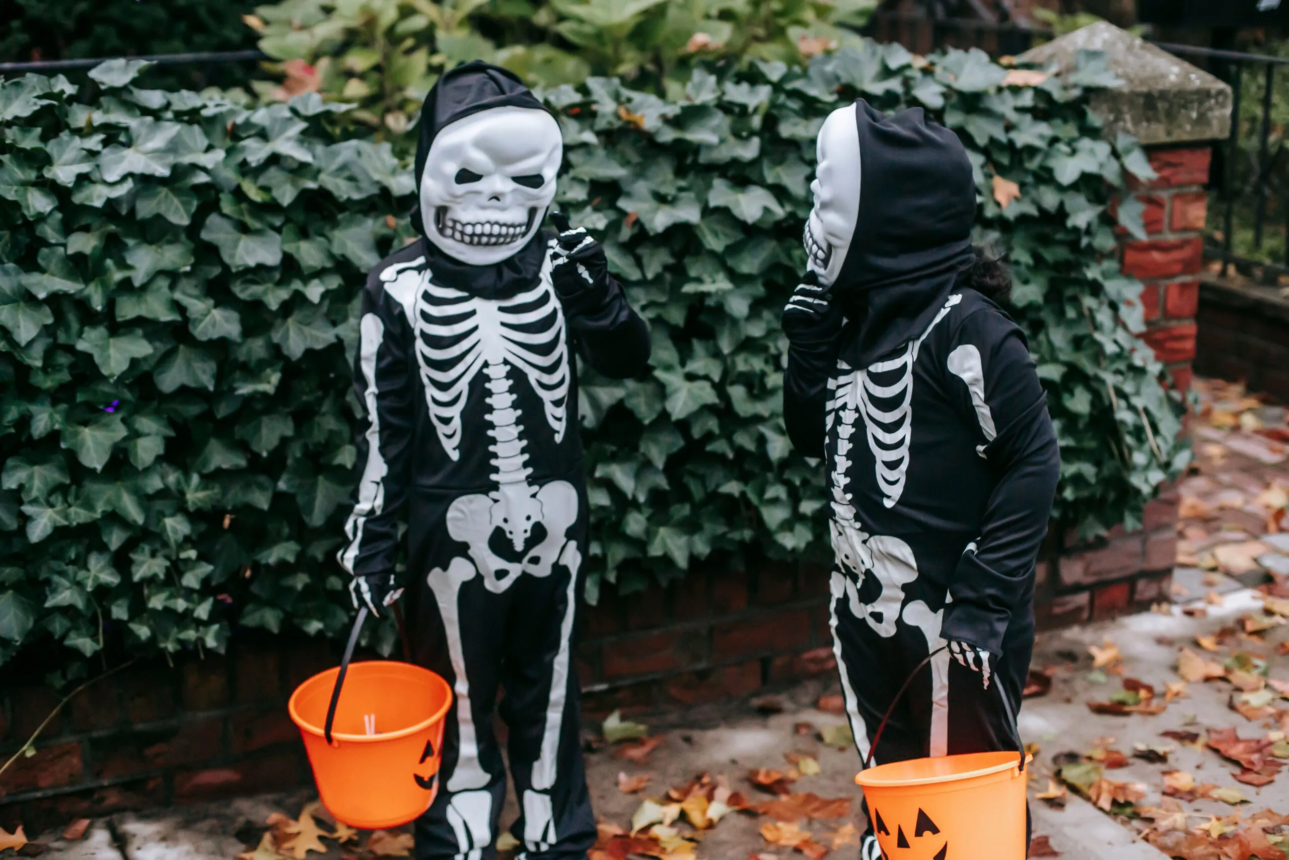 Anonymous kids wearing masks and skeleton costumes standing on street near green bush with orange buckets for trick or treat game