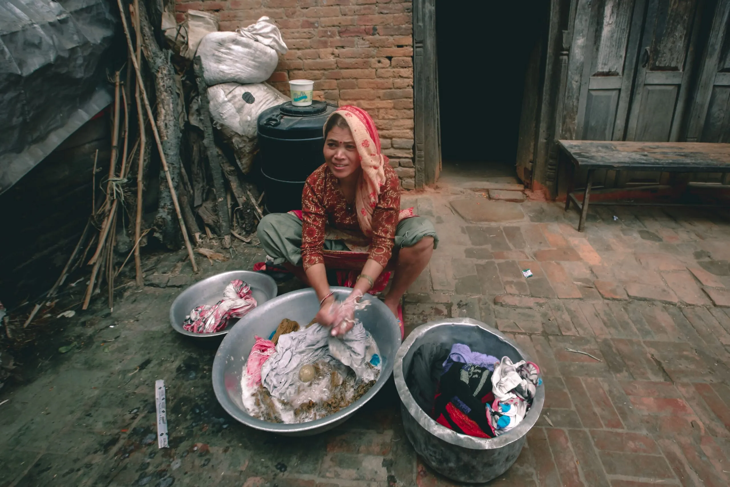 From above of content young ethnic female in traditional wear washing clothes in metal basins while looking away against old house
