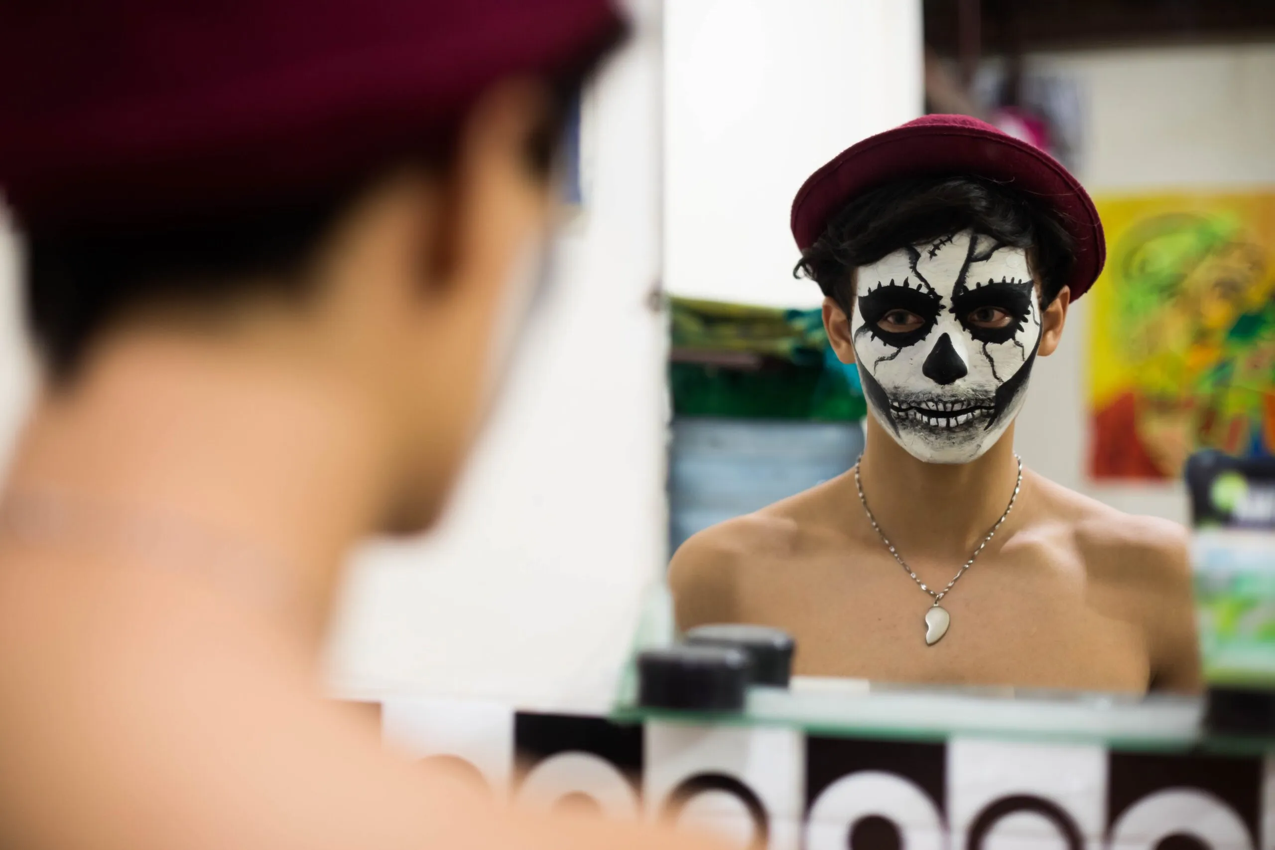 Young man with skull face paint and red hat looks into a mirror indoors.