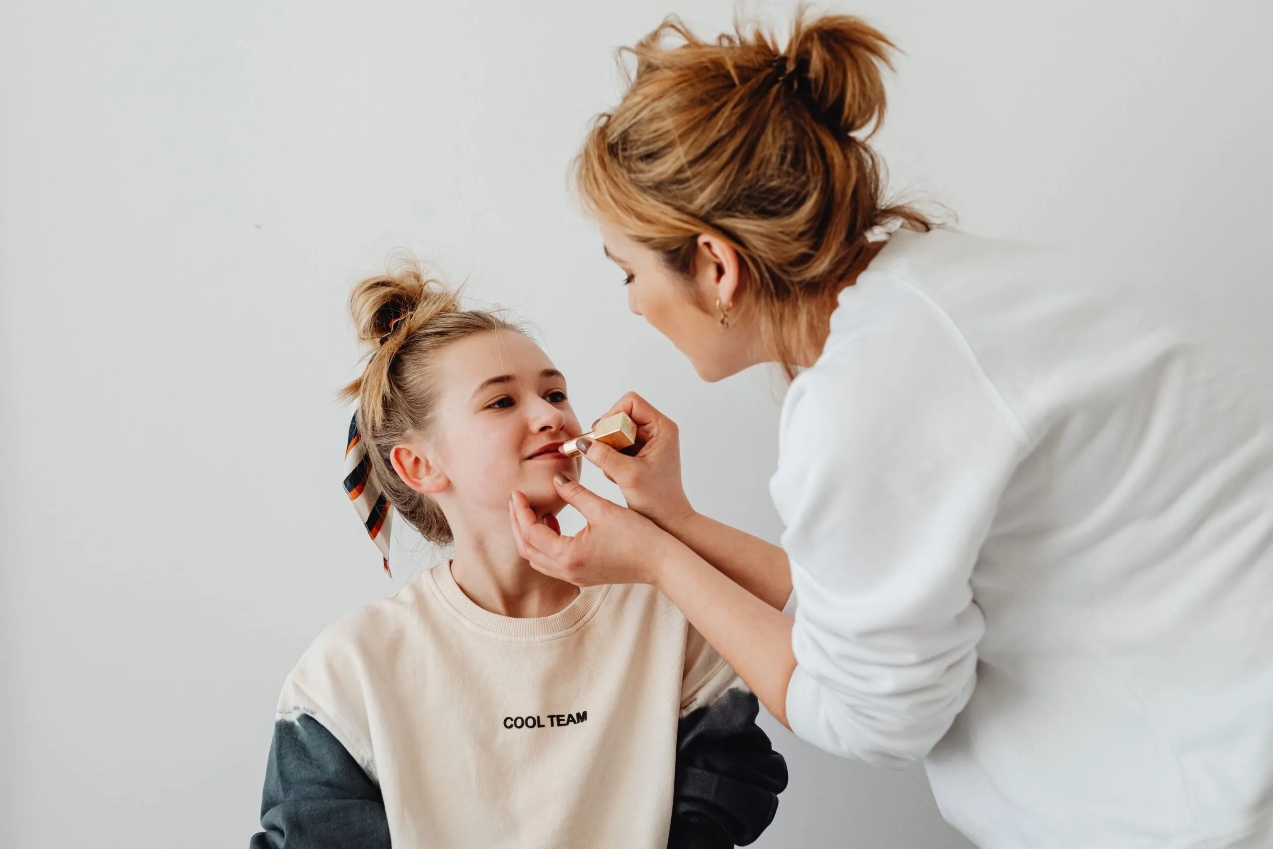 A mother lovingly applies lipstick to her daughter's lips, bonding over makeup indoors.