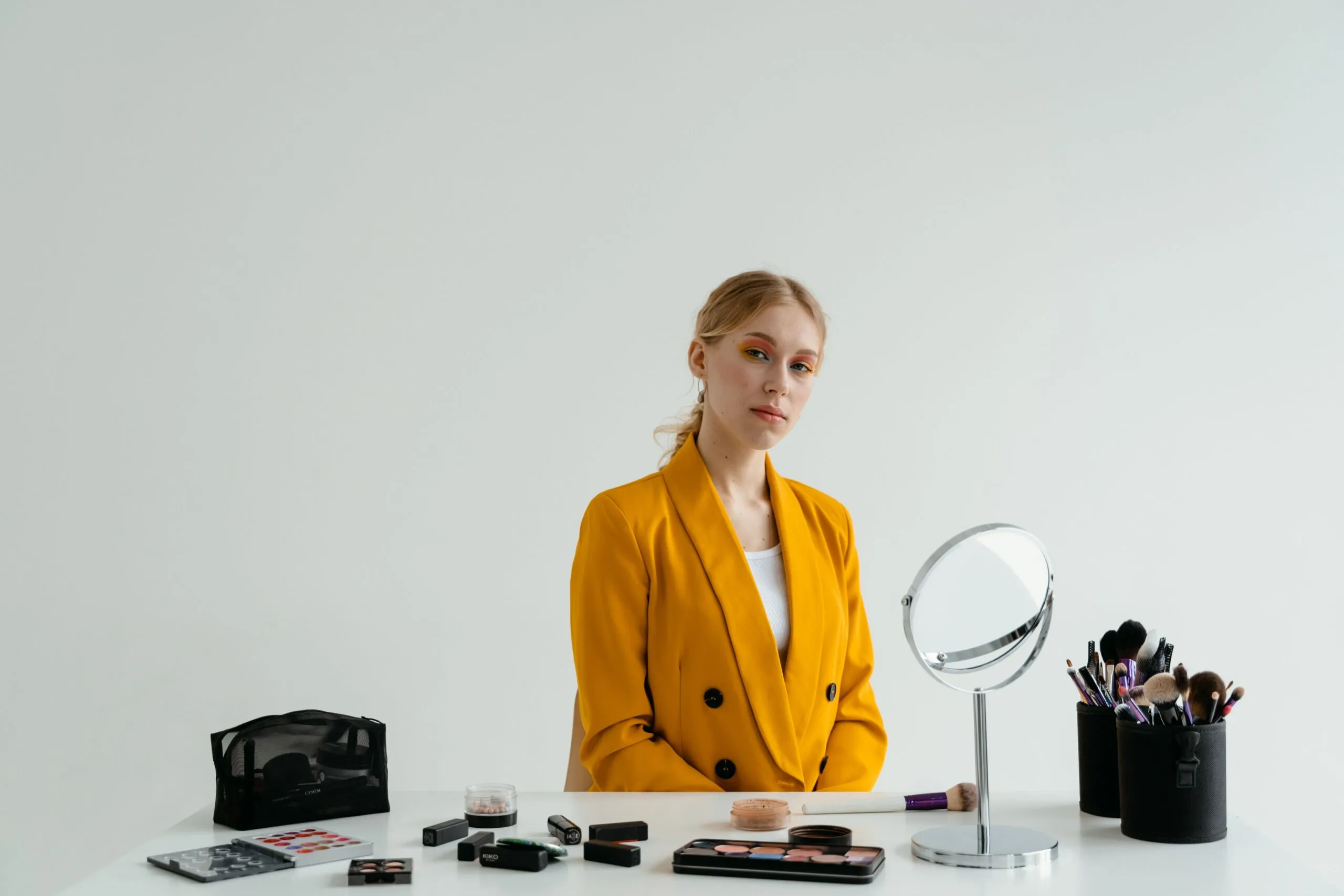 A woman in a yellow blazer sitting at a table with makeup tools and a mirror, ready for application.