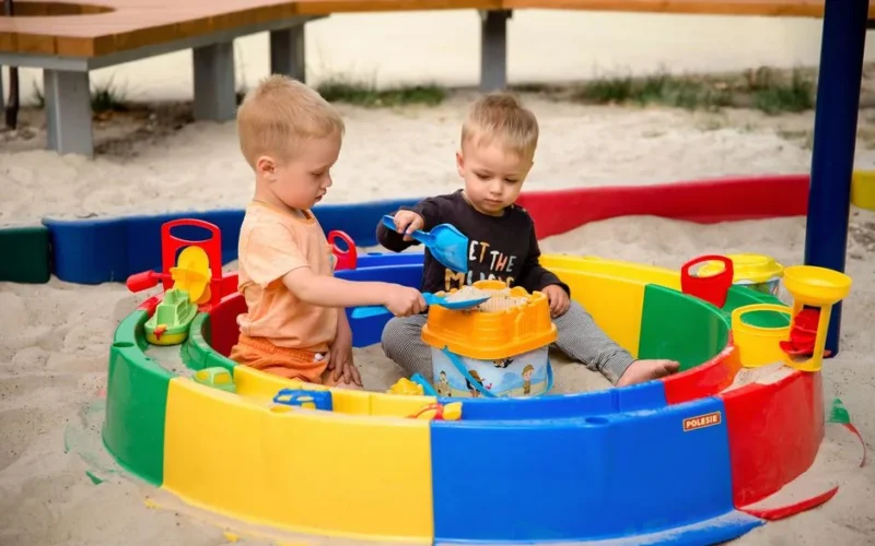 Two young children playing with toys in a colorful outdoor sandpit on a sunny day.