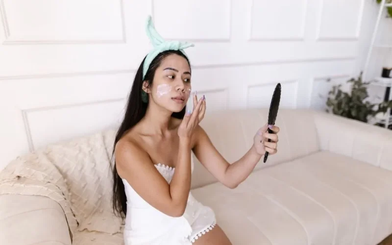 Asian woman in a towel applying face cream while looking in a handheld mirror indoors.