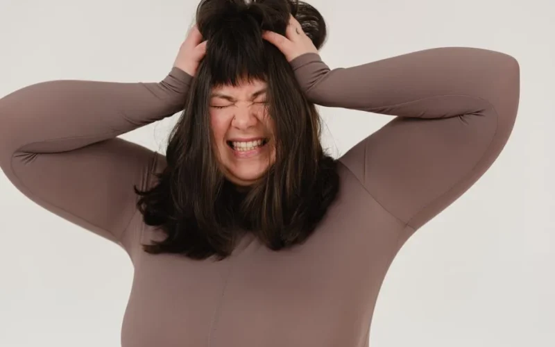 Expressive young overweight lady with dark hair grabbing head and shouting with closed eyes against white background