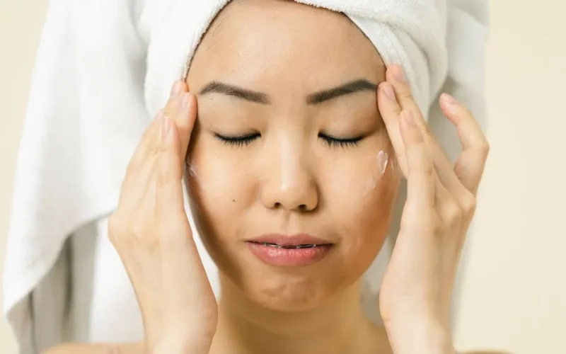 Asian woman with a towel on head applying moisturizer for skincare routine.