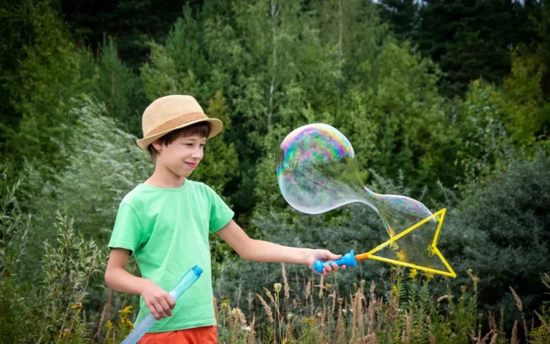 A young boy in a straw hat blows a giant bubble outdoors surrounded by lush greenery.