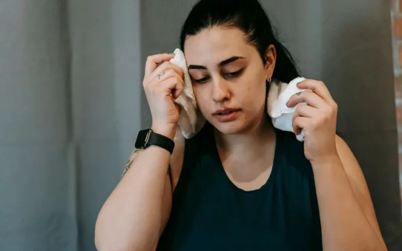 Exhausted overweight ethnic female with black hair in activewear wiping sweat from face looking down while training in modern fitness center