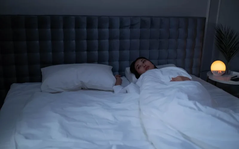 An Asian woman lying awake under soft white bedding in a dimly lit modern bedroom.