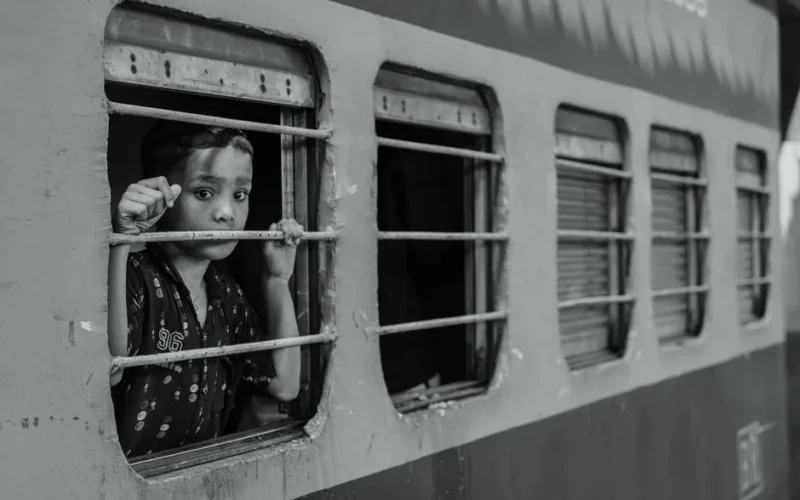 Black and white photo of a child looking pensively out of a train window, conveying emotion.