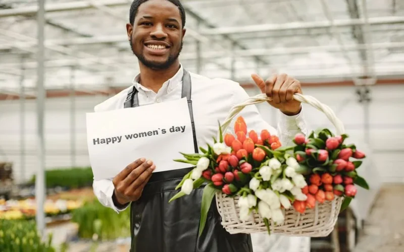A cheerful gardener holding a basket of colorful tulips and a Happy Women's Day sign in a greenhouse.