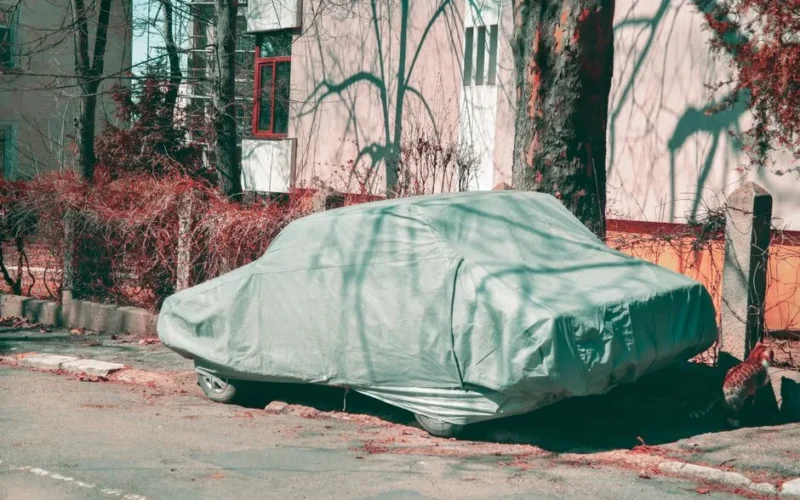 A covered car parked on an urban street surrounded by trees and buildings, casting shadows.