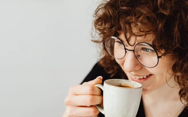 Smiling woman with curly hair and glasses savoring coffee in a cozy indoor setting.